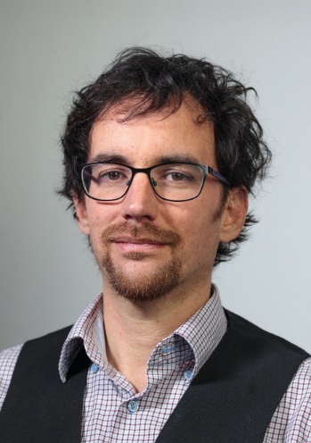 A person with glasses and beard wearing a vest and vest Description automatically generated